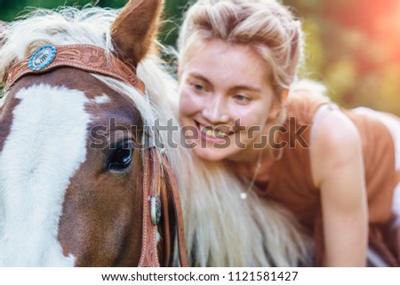 Portrait of happy smiling woman cowgirl, riding a brown horse. Clothed white jeans shorts, brown leather vest. Has slim sport body. Portrait nature. People and animals friendship concept.