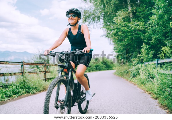 Portrait of a\
happy smiling woman dressed in cycling clothes, helmet and\
sunglasses riding a bicycle on the asphalt out-of-town bicycle\
path. Active sporty people concept\
image.