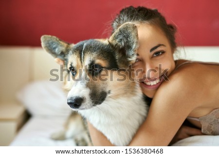 Portrait of happy smiling woman with her cute Welsh Corgi dog lying on the bed at morning, morning greeting. Beautiful brunette female hugging funny puppy, has good relationships with pet.