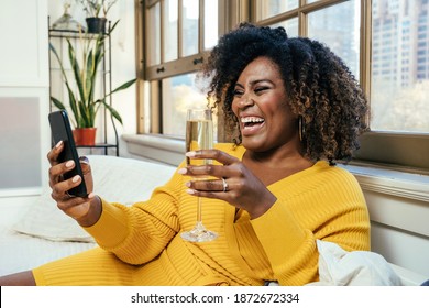 Portrait of a  happy smiling  woman laughing while  celebrating and talking with Champagne glass on a phone video call at home on couch