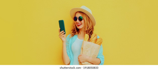 Portrait of happy smiling woman with phone holding grocery shopping paper bag with long white bread baguette on a yellow background