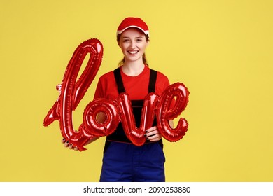 Portrait Of Happy Smiling Worker Woman Standing Holding World Love From Foil Balloon, Looking At Camera, Wearing Overalls And Red Cap. Indoor Studio Shot Isolated On Yellow Background.