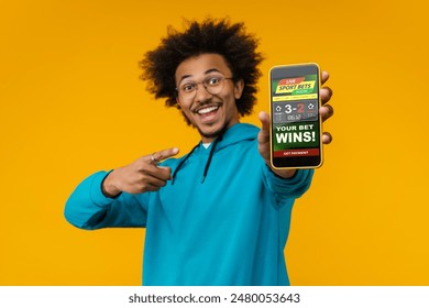 Portrait of happy smiling young African American man reaching smartphone with bookmaker's application interface on its screen. Sports bet, money win concept. Focus is in phone. - Powered by Shutterstock