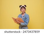 Portrait of happy smiling young Asian college student with notebook writing something ready to school isolated on yellow background.