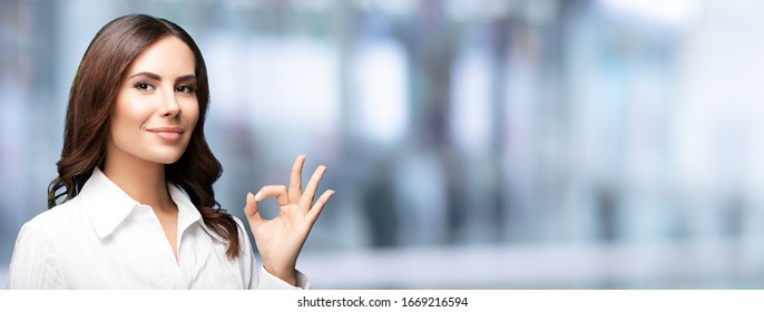 Portrait of happy smiling young businesswoman, showing ok hand sign gesture, with blank copy space for slogan or some text, over blurred office background. Success in business concept. 