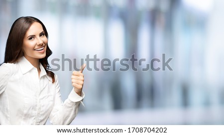 Portrait of happy smiling young excited businesswoman, showing thumb up hand sign gesture. Success in business concept studio shot. Standing against blurred office background.