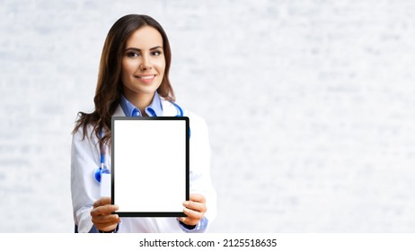Portrait of happy smiling young female doctor showing tablet pc ipad touchpad with mockup empty copy space area, over office white brick loft wall. Selective focus on mock up tablet pc. 