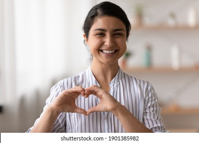 Portrait of happy smiling young lady of indian ethnicity looking at camera holding fingers joined in tender heart close to chest. Young woman volunteer video blogger ask audience to support charities