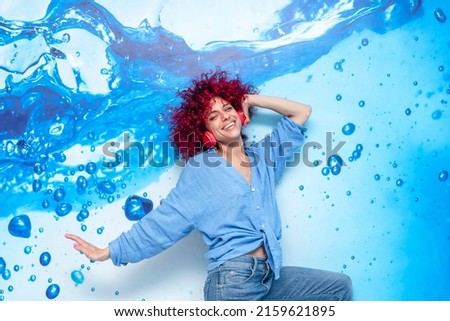 portrait of a happy smiling young latin woman with red afro hair having a good time dancing and listening to music and on her red headphones on a blue background with blue drops