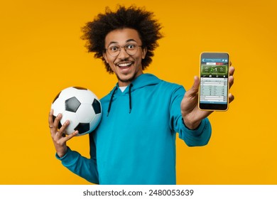 Portrait of happy smiling young man holding soccer ball in hand and reaching smartphone with bookmaker's application interface on its screen. Sport bet, money win concept. Focus is in phone. - Powered by Shutterstock