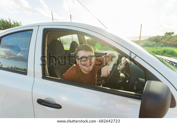 Portrait of happy smiling young man,\
buyer sitting in his new car and showing keys outside dealer\
office. Personal transportation, auto purchase\
concept