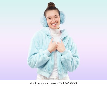 Portrait Of Happy Smiling Young Woman In Blue Earmuffs And Furry Blue Sweatshirt. Attractive Female Stands On Green Gradient Background, Front View. Concept Of Stylish Warm Clothes In Winter Time.