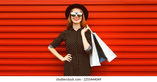Portrait of happy smiling young woman with shopping bags wearing black round hat on red background - Shutterstock ID 2255444049