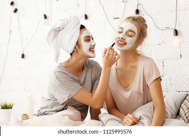 Portrait of happy smiling young women sitting in light bedroom and making fun using brush for applying facial cleansing mask. Home beauty treatments concept - Powered by Shutterstock