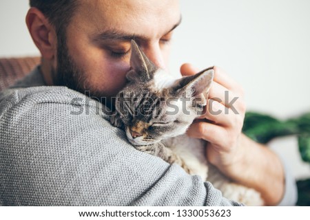 Portrait of happy snuggling cat with close eyes and young beard man. Handsome young guy is hugging and cuddling his cute color point Devon Rex Kitten. Domestic pets concept