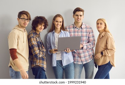 Portrait Of Happy IT Students With Laptop Computer Smiling And Looking At Camera Standing Together On Gray Studio Background. Group Of Young People Working On Project And Using Online Learning Tools