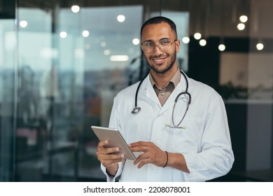 Portrait of happy and successful african american doctor man working inside office clinic holding tablet computer looking at camera and smiling wearing white coat with stethoscope - Powered by Shutterstock