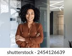 Portrait of happy and successful business woman, boss in shirt smiling and looking at camera inside office with crossed arms, african american woman with curly hair in corridor.