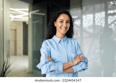 Portrait of happy and successful business woman, boss in shirt smiling and looking at camera inside office with crossed arms, Hispanic woman with curly hair in corridor. - Powered by Shutterstock