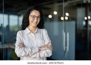 Portrait of happy and successful business woman, boss in shirt smiling and looking at camera inside office with crossed arms, Hispanic woman with curly hair. - Powered by Shutterstock