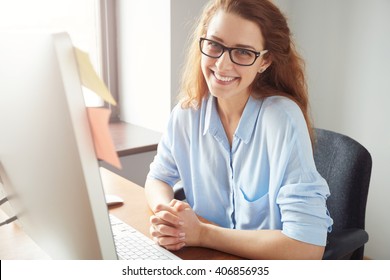 Portrait of happy successful Caucasian businesswoman in blue shirt and glasses looking and smiling at the camera. Attractive female manager sitting at desk after hard working day 