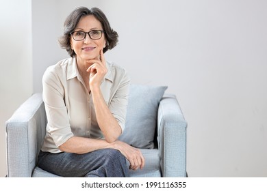 Portrait of happy, successful elderly woman with glasses sitting in a chair, smiling aged mature woman in stylish clothes looking at camera