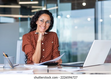 Portrait of happy and successful hispanic woman, businesswoman smiling and looking at camera holding contracts and invoices, working inside office with laptop on paper work. - Shutterstock ID 2226596727