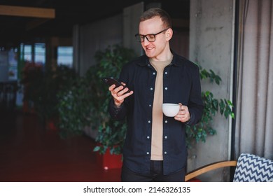 Portrait of a happy successful man in optical glasses holding a modern mobile phone and coffee in his hands, a cheerful blogger with a digital smartphone posing in a cafe