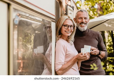 Portrait of happy successful mature couple spouses husband and wife smiling at the camera holding cups of coffee near their van camper wheel caravan home trailer - Powered by Shutterstock