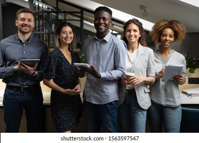 Portrait of happy successful multiracial business team standing with digital tablets, notebooks, ready to make notes. Smiling young motivated startup international employees group looking at camera.