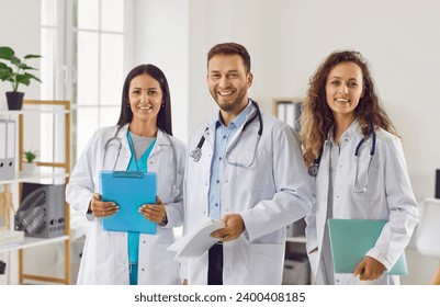 Portrait of a happy successful team of three young doctors in white uniform looking cheerful at camera and smiling while standing in hospital or clinic. Medical staff and health care concept. - Powered by Shutterstock