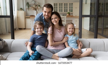Portrait Of Happy Successful Young Family With Children First Time Home Buyers Sit On Comfortable Couch, Smiling Parents And Little Kids Relax In Living Room Look At Camera Posing, Ownership Concept