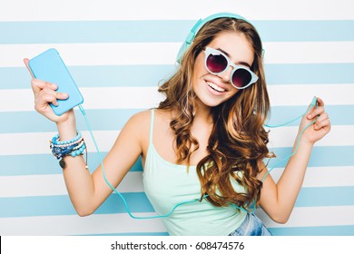 Portrait happy summer mood of joyful young woman with long curly hair, in sunglasses, heels having fun on striped background.Blue colors, expressing positivity, music, joy, happiness, dancing, smiling