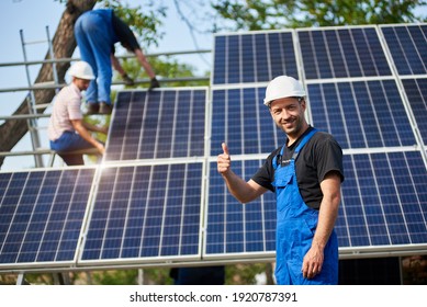 Portrait des glücklichen Technikers zeigt Daumen-up Geste stehen vor unfertigen High-Extra-Fotovoltaik-System blauen glänzenden Oberfläche mit Team von Mitarbeitern auf hoher Plattform.