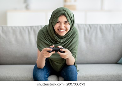Portrait Of Happy Teen Girl In Hijab Holding Joystick, Playing Videogame At Home. Positive Adolescent In Arabic Headscarf Engaged In Interesting Computer Game. Lockdown Pastimes And Hobbies