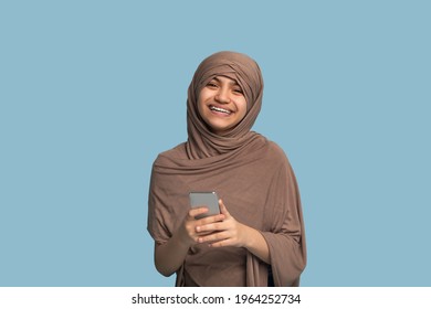 Portrait Of Happy Teen Girl In Islamic Hijab Using Smartphone Over Blue Studio Background. Adolescent In Traditional Muslim Headscarf Chatting Online, Posting In Social Media, Having Video Call