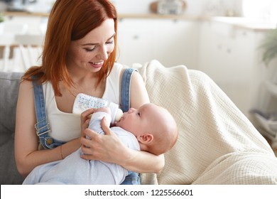 Portrait Of Happy Tender Young Mother With Ginger Hair Feeding Her Baby Boy From Milk Bottle. Beautiful Mom Feeds Infant Son With Formula While Sitting On Couch At Home. Motherhood And Happiness