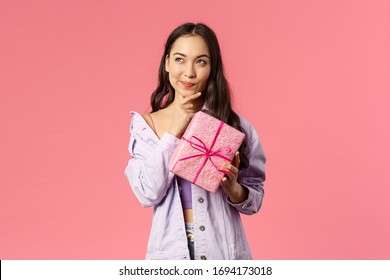 Portrait Of Happy Thoughtful Young Asian Girl Receive Wrapped Box With Gift, Wonder What Hidden Inside, Trying To Guess, Smile And Look Up Thinking, Standing Pink Background Intrigued