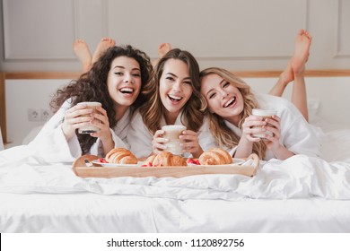 Portrait of happy three women 20s wearing bathrobe drinking latte and eating croissants in bed during hen party in posh apartment or hotel room