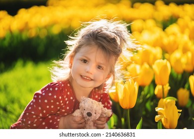 Portrait Of A Happy Three Year Old Girl With A Teddy Bear In Her Hands Against The Backdrop Of Flowers. Peace In Ukraine. No War.