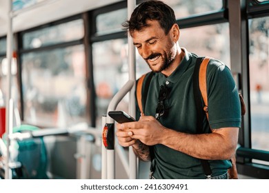 Portrait of a happy trendy guy commuting by public bus and using his cellphone while smiling at it.