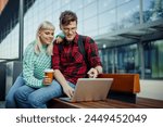 Portrait of a happy university students sitting on a bench on city street and pointing at the laptop while smiling at it. A hipster couple is using e-learning apps on the street. Students studying.