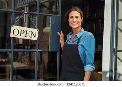 Portrait of a happy waitress standing at restaurant entrance. Portrait of mature business womanattend new customers in her coffee shop. Happy woman owner showing open sign in her small business shop. - Powered by Shutterstock