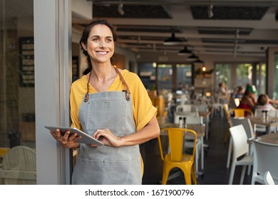 Portrait of a happy waitress standing at restaurant entrance holding digital tablet. Happy mature woman owner in grey apron standing at coffee shop entrance leaning while looking away with copy space. - Shutterstock ID 1671900796