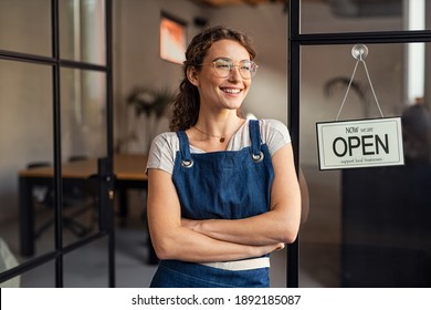 Portrait of happy waitress standing at restaurant entrance. Portrait of young business woman attend new customers in her coffee shop. Smiling small business owner showing open sign in her shop. - Shutterstock ID 1892185087
