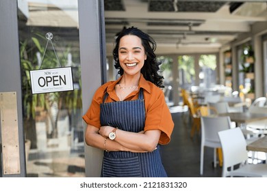 Portrait of happy waitress standing at restaurant entrance and looking at camera. Young business woman wearing apron standing with open sign at entrance gate while waiting for clients. 