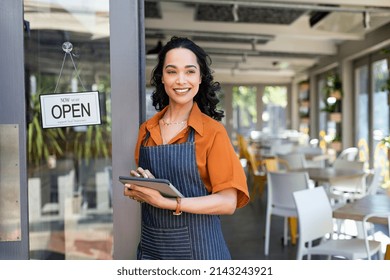 Portrait of happy waitress standing at restaurant entrance and holding digital tablet. Happy young latin woman owner in apron standing at coffee shop entrance, leaning while waiting clients.  - Powered by Shutterstock