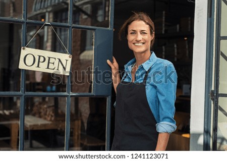 Portrait of a happy waitress standing at restaurant entrance. Portrait of mature business womanattend new customers in her coffee shop. Happy woman owner showing open sign in her small business shop.