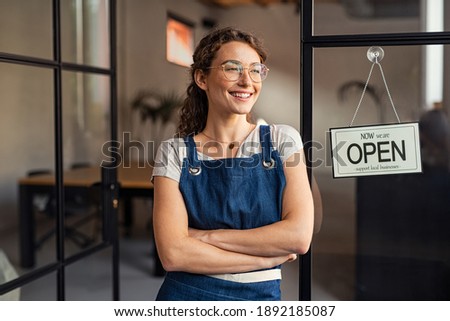 Portrait of happy waitress standing at restaurant entrance. Portrait of young business woman attend new customers in her coffee shop. Smiling small business owner showing open sign in her shop.