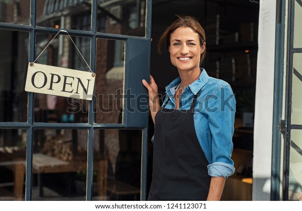 Portrait of a happy waitress standing at restaurant
entrance. Portrait of mature business womanattend new customers in
her coffee shop. Happy woman owner showing open sign in her small
business shop.